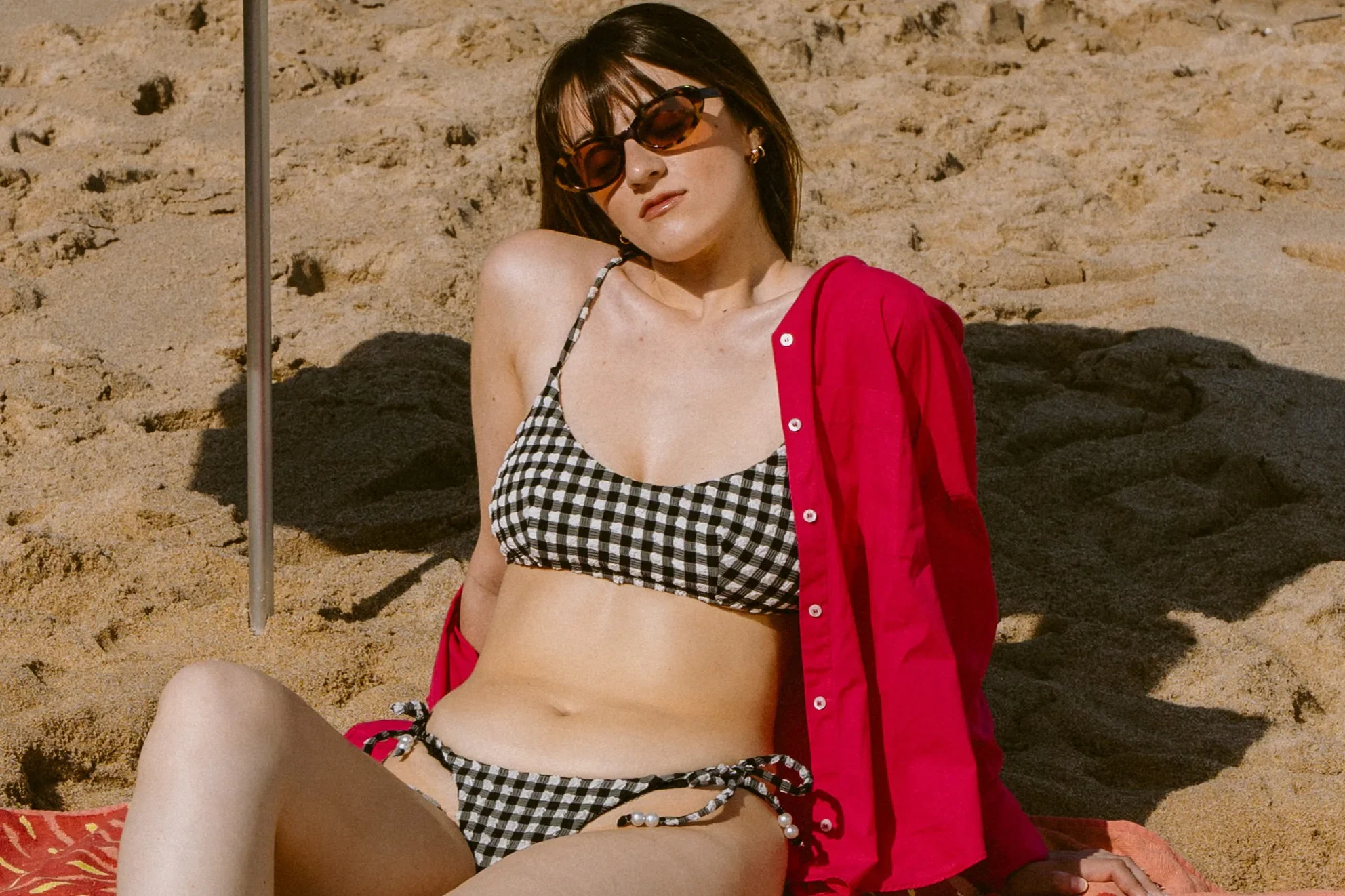 Une femme assise sur le sable en maillot de bain deux pièces en vichy noir et blanc, une veste rouge sur les épaules